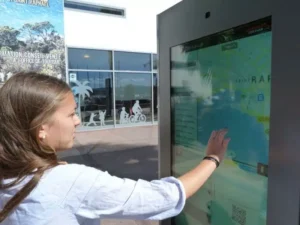 Voyageuse utilisant une borne digitale interactive devant un office de tourisme pour explorer une destination.